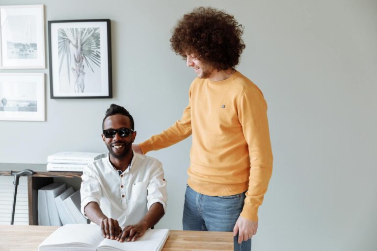 Two men at a table, one uses braille, while the other offers support. Indoors setting.